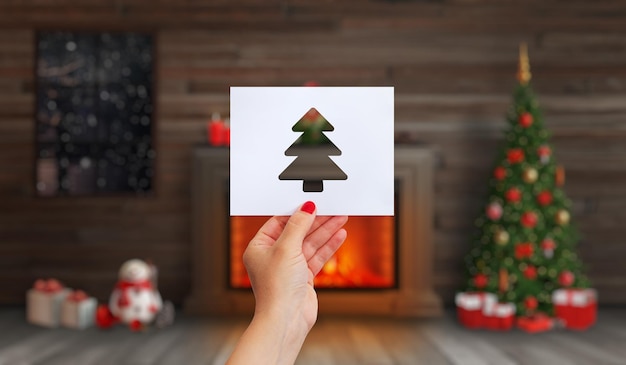 La mano tiene una carta con un albero di Natale tagliato su di esso Camino con un fuoco un albero di Natale e decorazioni natalizie