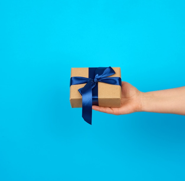 La mano tiene una scatola con un regalo avvolto in carta kraft marrone e legato con un nastro di seta blu