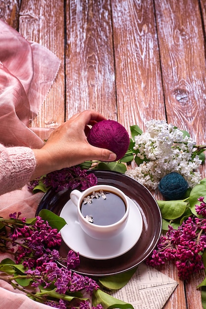 La mano tiene una tazza di caffè mattutino con rami di fiori lilla primaverili che sbocciano su sfondo di legno vista dall'alto. Stile sotterraneo piatto. Colori costosi. Design creativo di fiori.