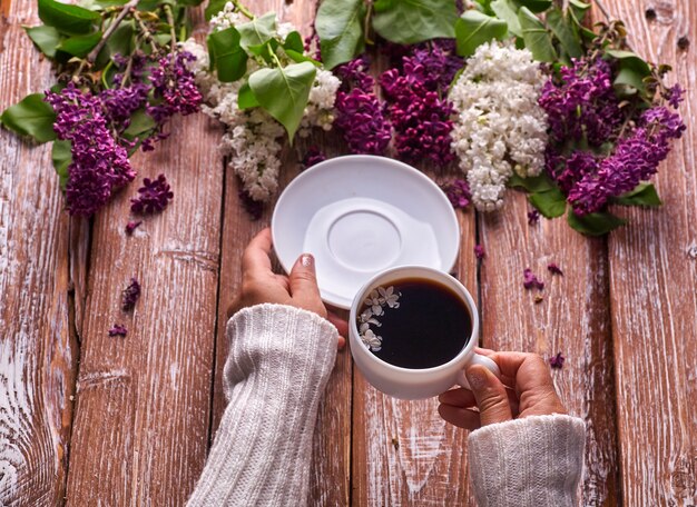 La mano tiene una tazza di caffè mattutino con rami di fiori lilla primaverili che sbocciano su sfondo di legno vista dall'alto. Stile sotterraneo piatto. Colori costosi. Design creativo di fiori.