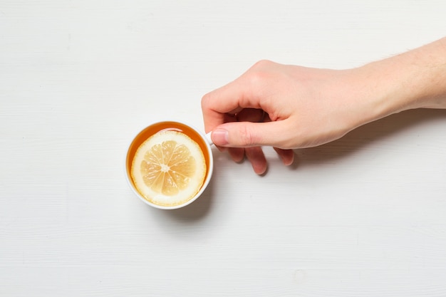 La mano tiene una tazza di tè al limone su un fondo di legno bianco. Vista dall'alto.