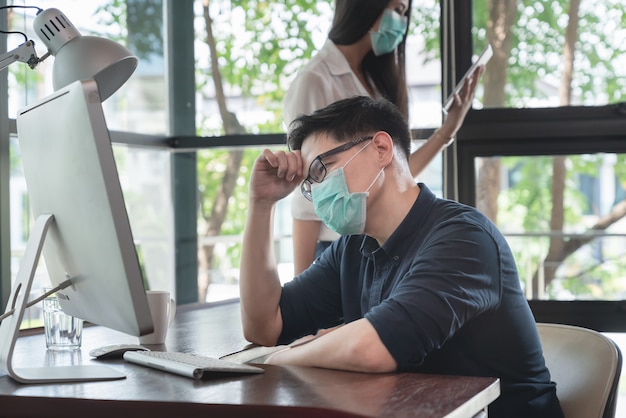 La maschera di protezione d'uso del giovane uomo creativo asiatico sollecitato per protegge il coronavirus che lavora all'ufficio del posto di lavoro