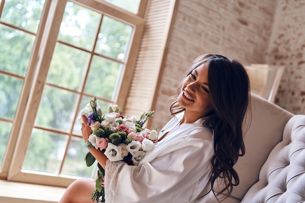 La mattina inizia con i fiori. Attraente giovane donna in accappatoio di seta con in mano un bouquet e sorridente mentre è seduta sul divano