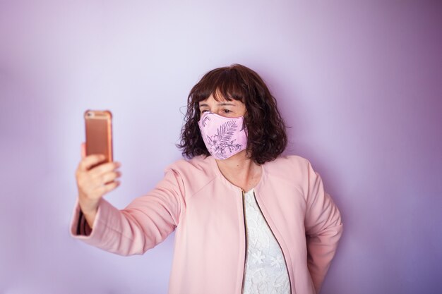 La mezza età capelli ricci donna che indossa rosa maschera medica utilizzando un telefono cellulare su uno sfondo rosa