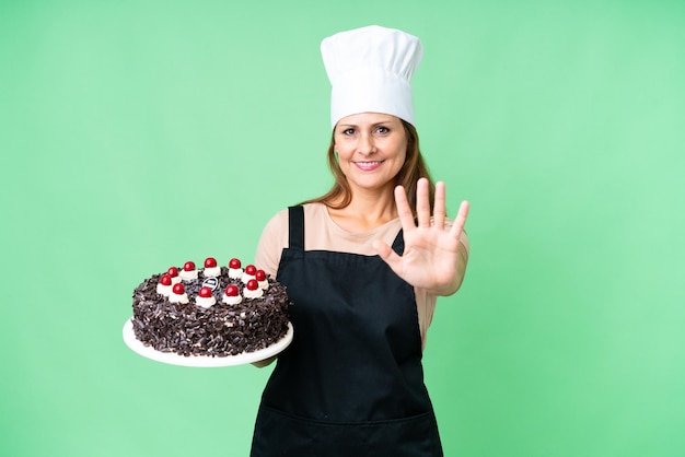 La mezza età pasticcere donna tenendo una grande torta su sfondo isolato contando cinque con le dita