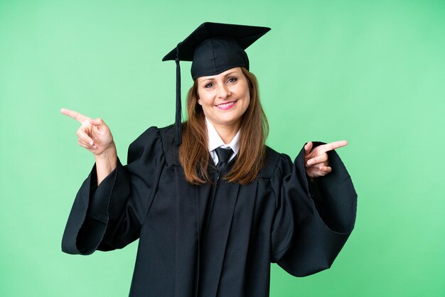 La mezza età universitaria donna laureata su sfondo isolato puntare il dito verso i laterali e felice