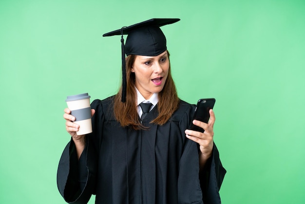 La mezza età universitaria donna laureata su sfondo isolato tenendo il caffè da portare via e un cellulare