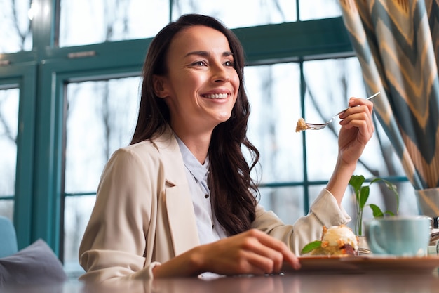 La mia debolezza. Elegante donna soddisfatta sognante degustazione di pasti seduti vicino alla finestra e sorridendo