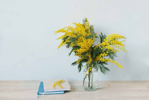 La mimosa si ramifica in un vaso di vetro sul tavolo un diario e un taccuino una parete blu come sfondo