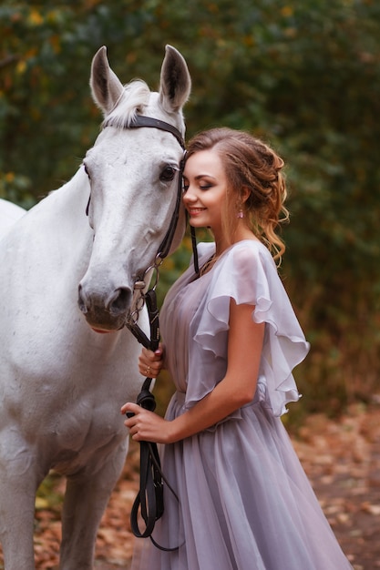 la modella in abito sorride e tiene in mano un cavallo bianco. Sfondo sfocato, effetto artistico. sfondo verde frondoso