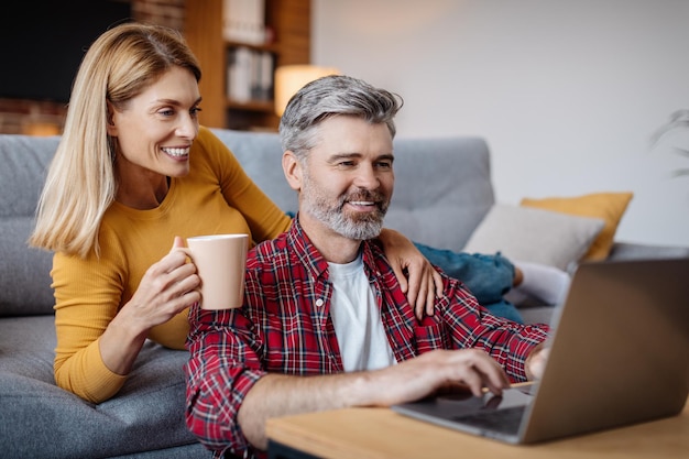 La moglie e il marito caucasici maturi felici che guardano il computer portatile si godono la pausa caffè nel tempo libero nel soggiorno