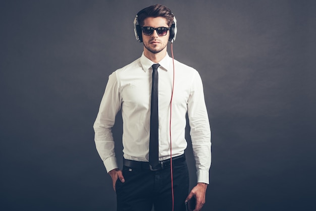 La musica è il suo stile. Bel giovane ben vestito in cuffia che guarda l'obbiettivo mentre sta in piedi su sfondo grigio