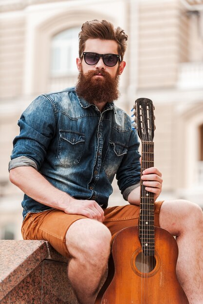 La musica è la mia vita. Fiducioso giovane uomo barbuto che tiene la chitarra e guarda la telecamera mentre è seduto all'aperto