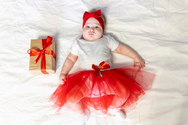 La neonata con una gonna rossa e un fiocco rosso in testa giace sul letto con un regalo di natale con rosso