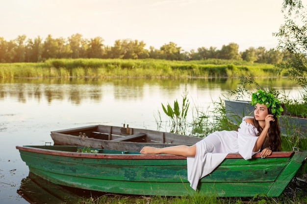 La ninfa dai lunghi capelli scuri in un abito bianco vintage seduta su una barca in mezzo al fiume. Tra i capelli una corona di foglie verdi con fiori bianchi. Servizio fotografico di fantasia.