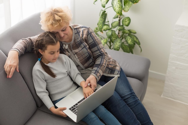 La nipotina intelligente della bambina si siede sul divano mentre fa shopping online con la nonna anziana eccitata, la nipote adolescente e la nonna sorridente comprano sul computer portatile che naviga in internet rilassandosi a casa insieme