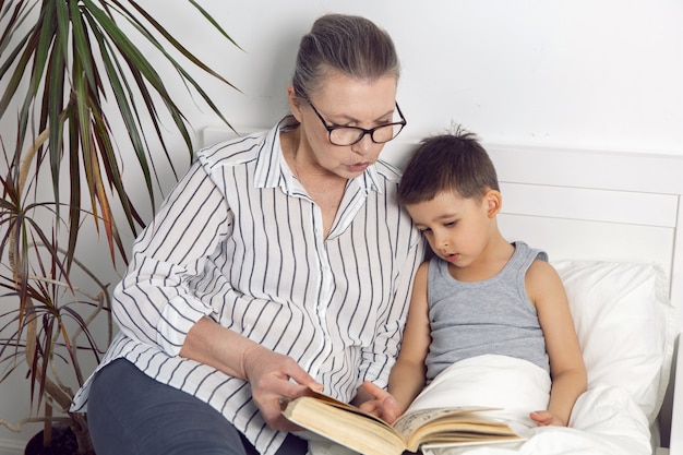 La nonna con gli occhiali e una camicia bianca legge un libro a suo nipote sdraiato sul letto