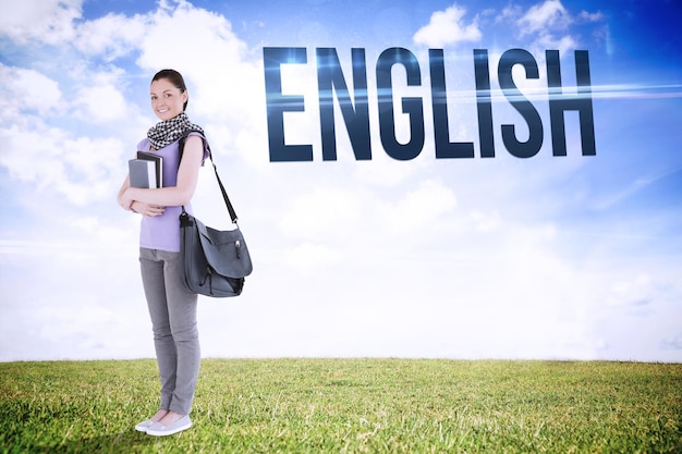 La parola inglese e studente attraente che tiene libri e la sua borsa mentre si trova contro un paesaggio sereno