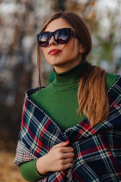 La passeggiata alla moda accogliente della ragazza al parco variopinto di autunno in occhiali da sole