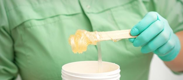 La pasta di zucchero gialla liquida per la depilazione su un bastoncino scorre nel barattolo nelle mani di un'estetista.