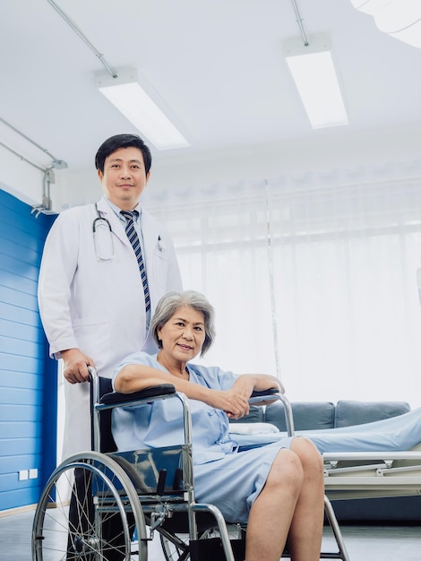 La paziente asiatica della donna anziana vestita di sorridere azzurro si siede felicemente sulla sedia a rotelle con il medico gentile dell'uomo in vestito bianco che sta accanto a lei vicino al letto nella stanza di risveglio nello stile verticale dell'ospedale