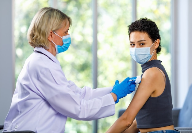 La paziente donna carina si siede in attesa di covid 19 vaccinando dall'ago della siringa dopo che la dottoressa caucasica in camice bianco con maschera facciale e guanti di gomma usa la spalla per la pulizia del cotone alcolico.