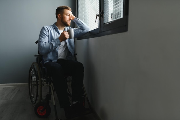 La persona disabile si siede in sedia a rotelle contro la finestra Uomo caucasico triste serio che indossa abiti casual e guarda un'ampia vista panoramica in un soggiorno o in un ospedale moderno e luminoso