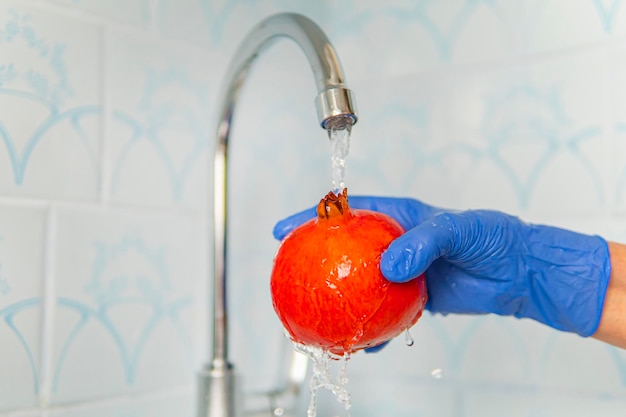 La persona lava la frutta con il sapone ecologico per il lavaggio in una cucina con pareti blu. Decontaminazione e disinfezione dei prodotti dal punto vendita. Prevenzione del coronavirus. le mani nei guanti lavano il granato rosso