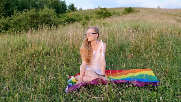 La persona omosessuale della donna lesbica bisessuale si siede sulla bandiera LGBT in bianco sulle verdi colline in una giornata di sole al tramonto e celebra una parata gay Giornata della bisessualità o Giornata nazionale del coming out nel mese dell'orgoglio