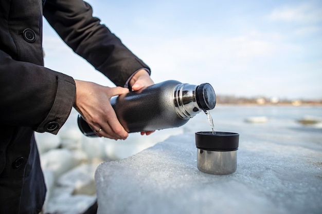 La persona versa il tè caldo da un thermos in una tazza che si trova su un lastrone di ghiaccio sulla riva del fiume