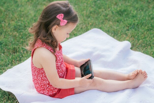 La piccola 3-4 ragazza in abiti rossi si siede su una coperta sull'erba verde e guarda nel telefono cellulare. Bambini, usando i gadget