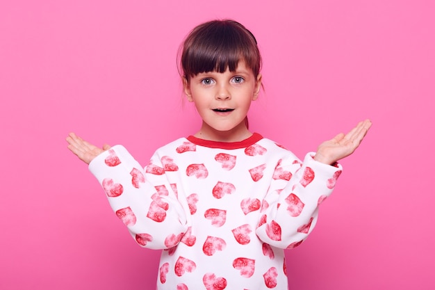 La piccola bambina sorpresa veste il maglione con i cuori, guardando la parte anteriore con la bocca aperta, ascoltando notizie scioccanti, allargando le mani da parte, isolato sopra il muro rosa