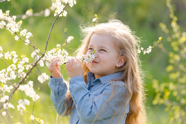 La piccola bionda carina con i capelli lunghi annusa un ramo di un albero in fiore nel parco in primavera.