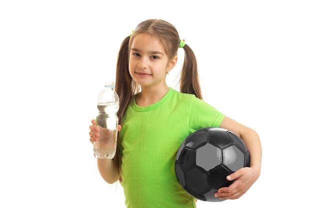La piccola donna sveglia con il pallone da calcio nelle mani beve l'acqua in bottiglia isolata su bianco