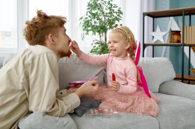 La piccola figlia che mette il rossetto sulle labbra di papà applicando cosmetici per il trucco si gode del tempo con i genitori a casa