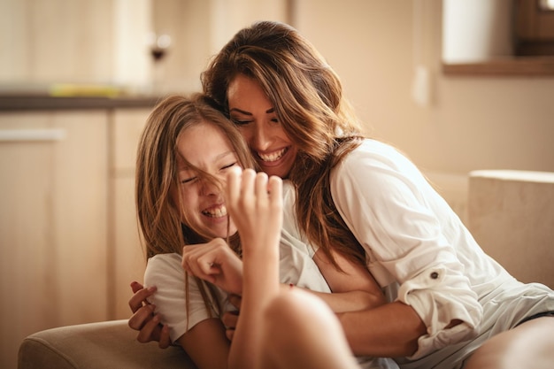 La piccola figlia e la sua giovane madre si divertono a farsi il solletico e a ridere abbracciandosi sul letto della loro casa.