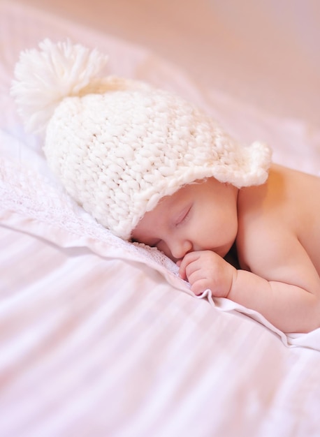 La piccola neonata indossa il cappello lavorato a maglia sdraiato a letto sopra il primo piano bianco. Infanzia.