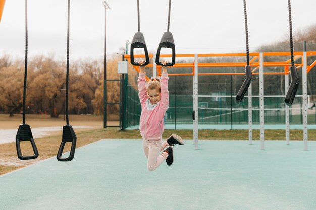 La piccola ragazza allegra gioca nel parco giochi. Il bambino è impegnato su anelli di ginnastica per strada. Il bambino pratica sport sui simulatori.