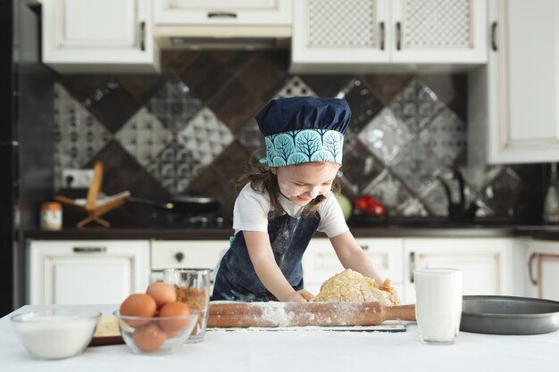 La piccola ragazza allegra impasta la pasta con le sue mani in cucina