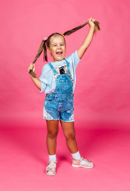 La piccola ragazza allegra in salopette di jeans tira i capelli in direzioni diverse. figli.