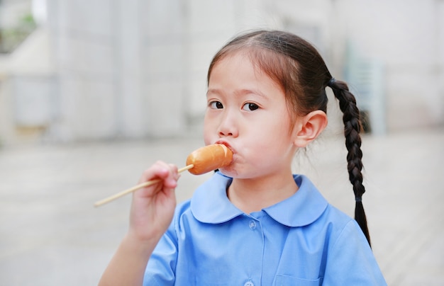 La piccola ragazza asiatica adorabile del bambino in uniforme scolastica gode di di mangiare la salsiccia.