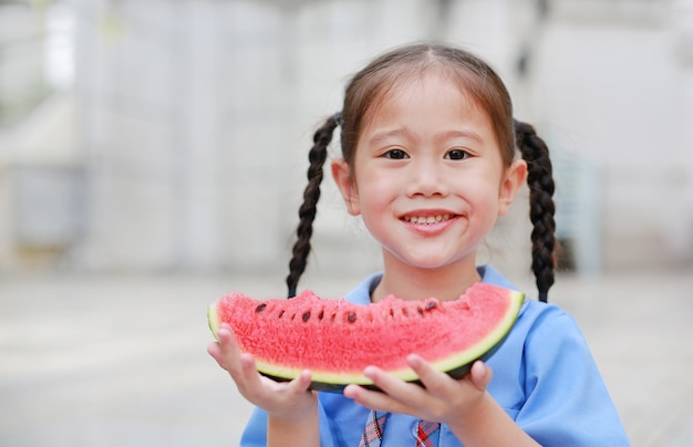 La piccola ragazza asiatica felice del bambino in uniforme scolastica gode di di mangiare l&#39;anguria all&#39;aperto.
