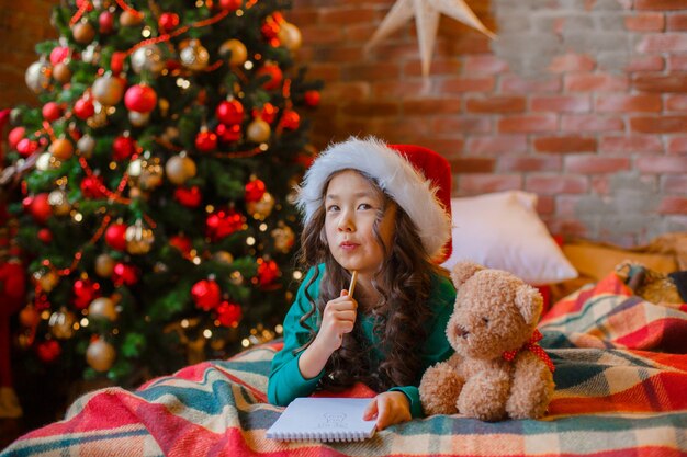 La piccola ragazza asiatica in pigiama vicino all'albero di Natale scrive una lettera al capodanno di Natale di Babbo Natale