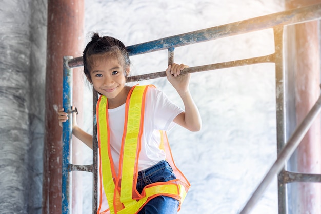 La piccola ragazza asiatica indossa una maglietta di sicurezza che gioca in cantiere. Apprendimento e concetto di lavoro da sogno.