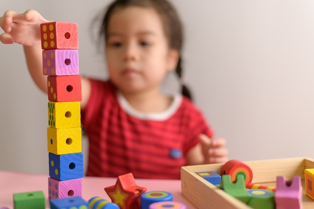 La piccola ragazza asiatica riccia gode di di giocare con i blocchetti di legno del giocattolo isolati sulla parete bianca. Istruzione e concetto di apprendimento.