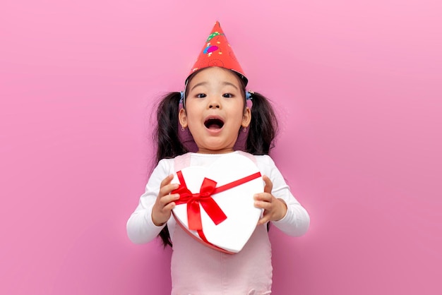 la piccola ragazza asiatica scioccata festeggia il compleanno con un berretto festivo e tiene il cuore regalo su sfondo rosa
