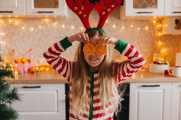 La piccola ragazza bionda carina sorridente in pigiama e corna tiene le metà della frutta vicino ai suoi occhi. bambino in cucina vicino all'albero di Natale. Natale