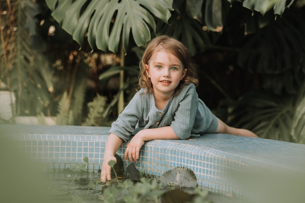 La piccola ragazza bionda è sdraiata sul lato di una piscina con piante acquatiche nel giardino botanico