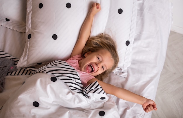 La piccola ragazza bionda felice in pigiama si estende a letto nella stanza