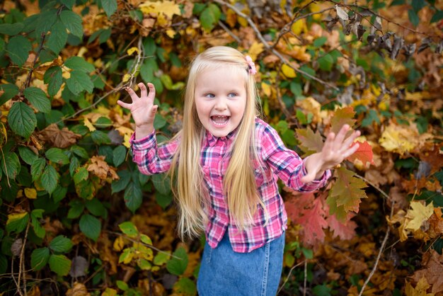 La piccola ragazza bionda negli shorts di una camicia e del denim cammina nel parco di autunno.